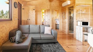 Ein gemütliches Holzhaus-Interieur mit einem grauen Ecksofa, einem Essbereich mit weißem Tisch und Stühlen sowie einer Küche mit einer Mikrowelle.