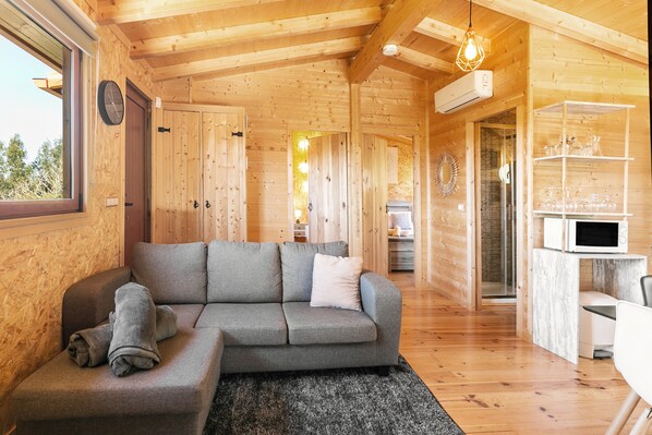 A cozy wooden cabin interior with a grey sectional sofa, a dining area with a white table and chairs, and a kitchen with a microwave.