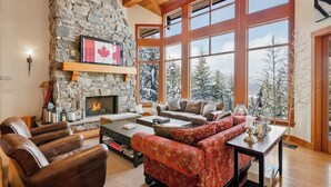 Una sala de estar acogedora con chimenea de piedra, sofás de cuero y ventanas amplias con vistas a árboles nevados.