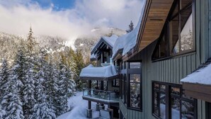 Un edificio moderno con balcón, rodeado de árboles y montañas cubiertas de nieve.