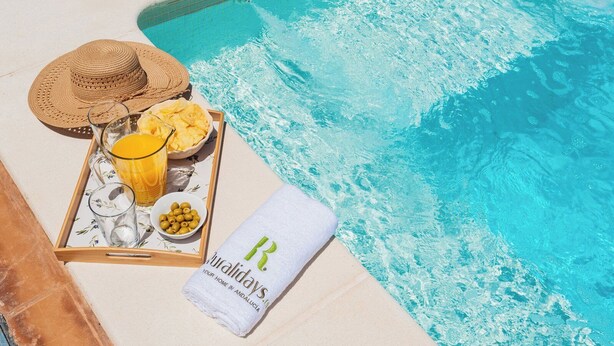 A beachside setting with a pool, a tray with drinks and snacks, a straw hat, and a towel with a logo. - Aromas de Alcala Alcalá La Real by Ruralidays (Jaén)