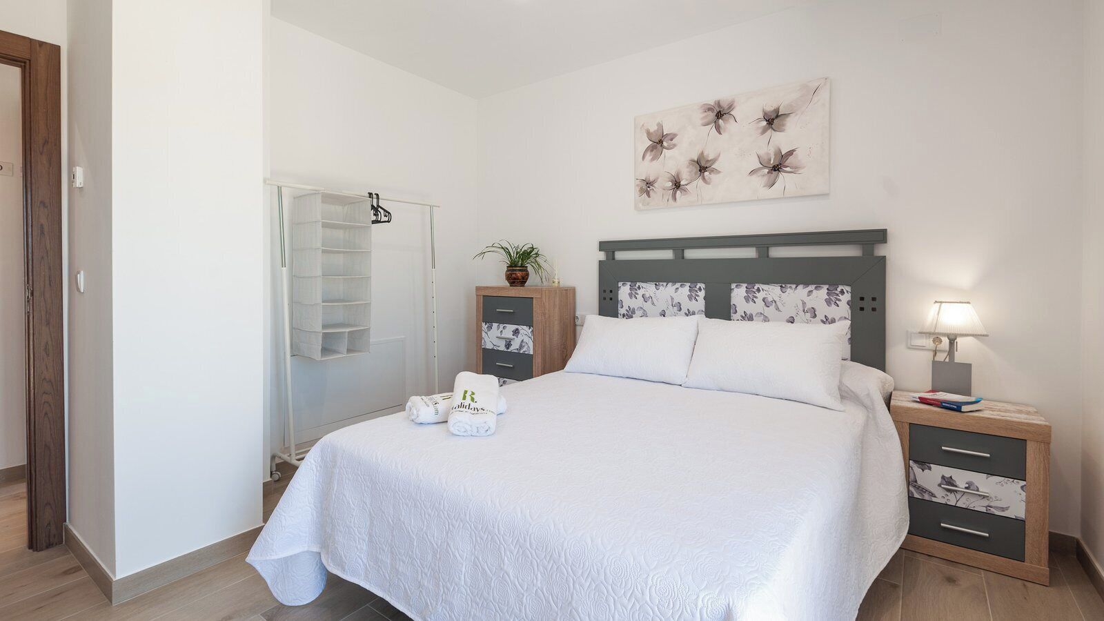 A neatly arranged bedroom with a bed, a nightstand, a lamp, a potted plant, and a wall-mounted artwork.