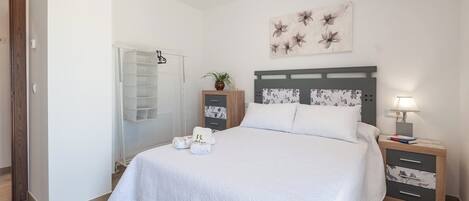 A neatly arranged bedroom with a bed, a nightstand, a lamp, a potted plant, and a wall-mounted artwork.