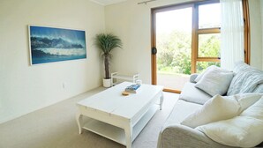Un salon avec une table basse blanche, un canapé gris agrémenté de coussins blancs, une plante en pot et une grande fenêtre donnant sur un paysage verdoyant.