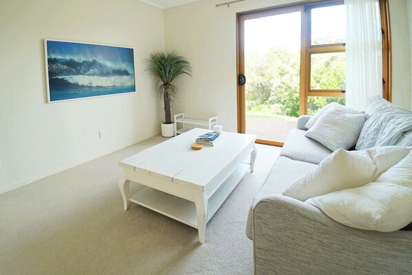 A living room with a white coffee table, a grey sofa with white cushions, a potted plant, and a large window with a view of greenery.
