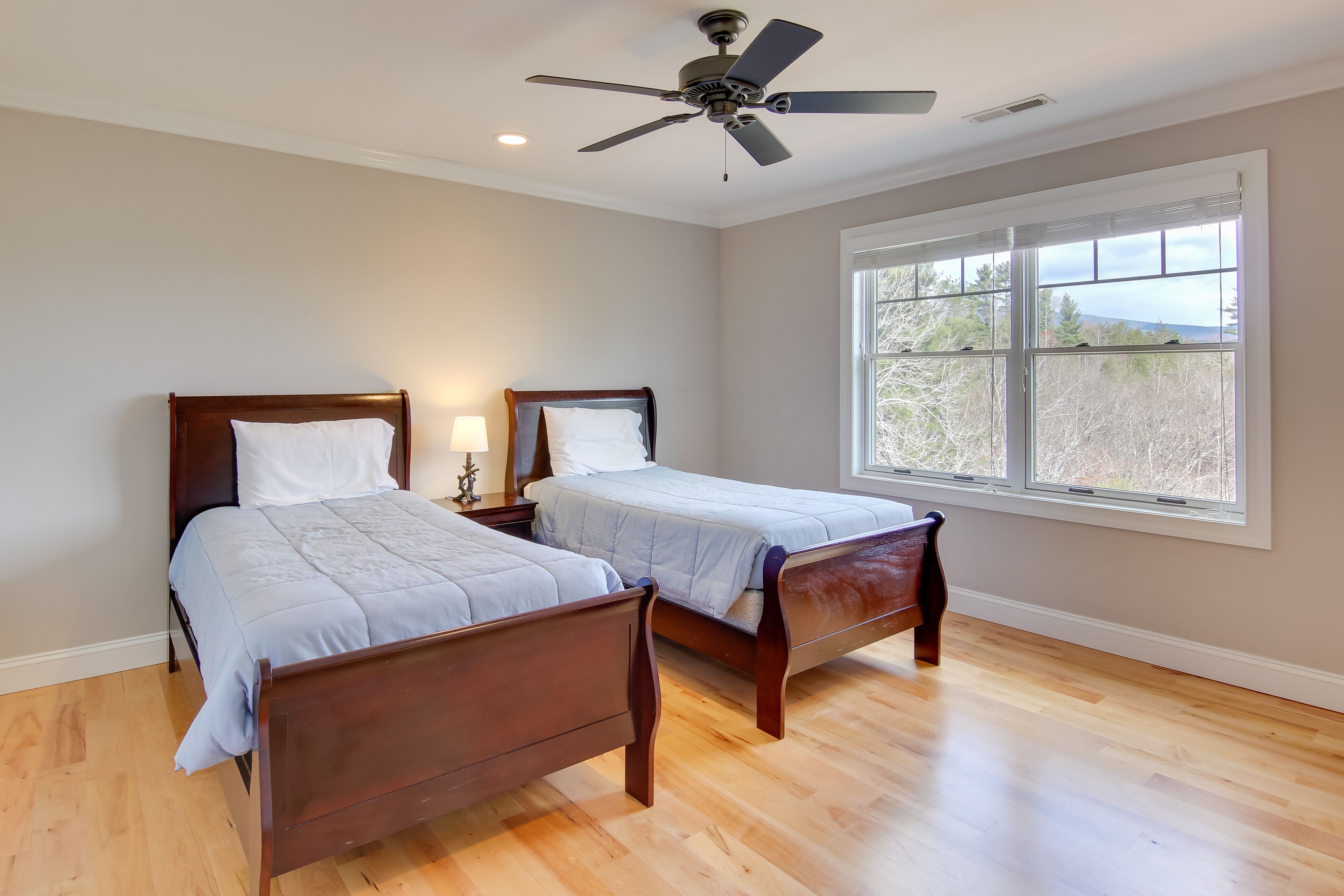 A bedroom with two wooden beds, a ceiling fan, and a large window with a view of trees.