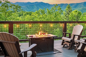 Une terrasse avec des fauteuils Adirondack, un barbecue où les flammes dansent, et une vue sur des montagnes verdoyantes.