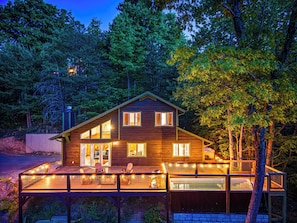 Une cabane en bois à deux étages, dotée d’une terrasse, de grandes fenêtres et d’un intérieur visible, avec une cheminée.