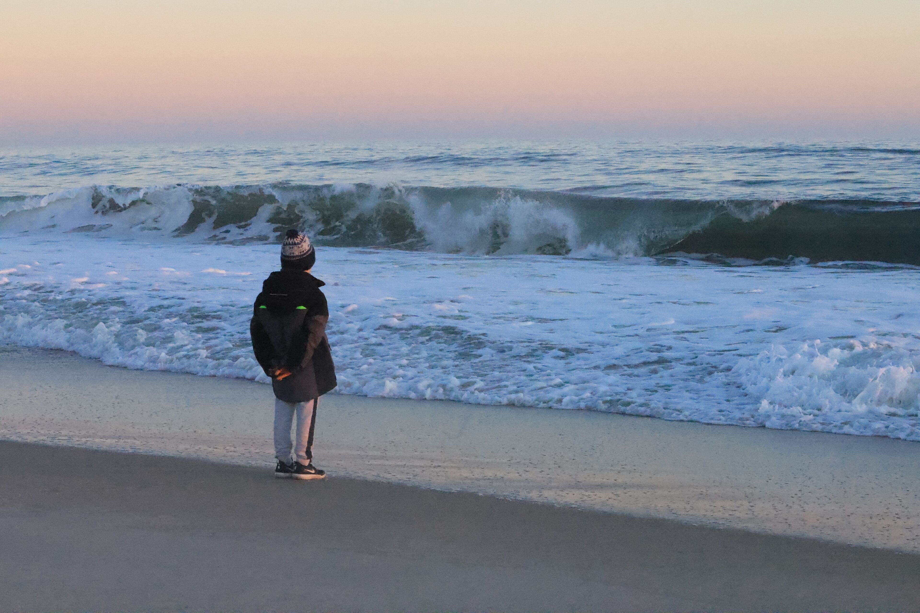 A person standing on a beach facing the ocean with waves crashing onto the shore.