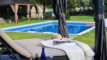 Una silla de descanso junto a la piscina con un libro y un vaso de agua, un pabellón al fondo y una vista despejada de la alberca.