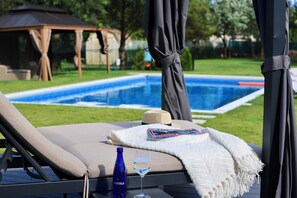 Une chaise longue placée au bord de la piscine, avec un livre et un verre d’eau à portée de main, un gazebo en arrière-plan et une vue dégagée sur la piscine.
