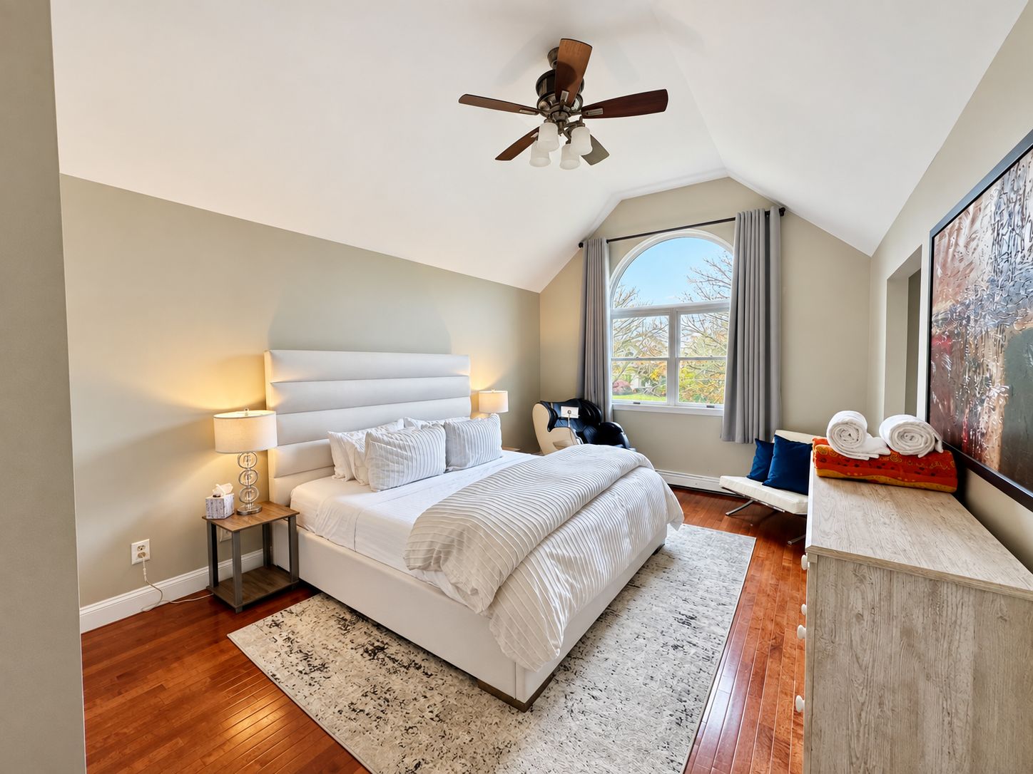 Une chambre à coucher comprenant un lit, des tables de chevet, un ventilateur de plafond, une fenêtre avec des rideaux et un grand tableau accroché au mur.