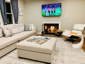 A living room with a beige sofa, a white armchair, a coffee table with books, a fireplace, and a TV displaying a soccer game.