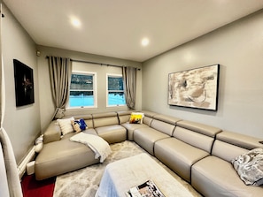 A modern living room with a beige sectional sofa, two windows with curtains, a wall-mounted abstract painting, and a red carpet.