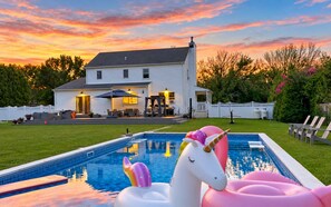 A swimming pool with inflatable unicorns, a white house with a porch, and a sunset sky.