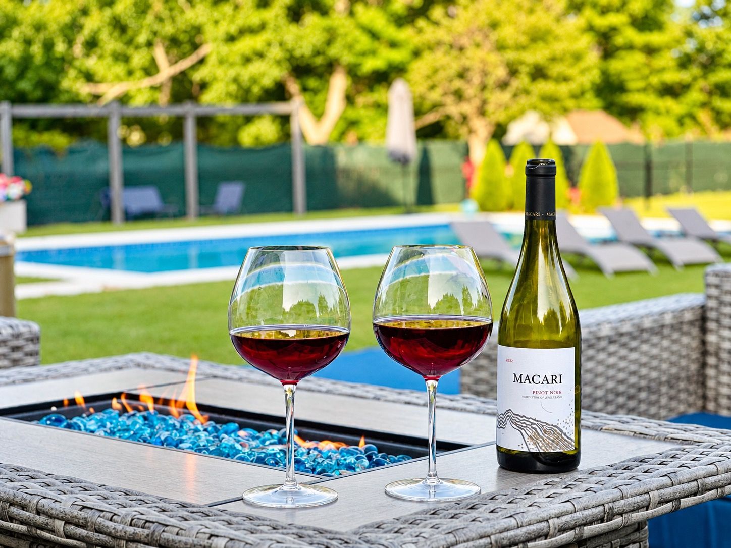 Une bouteille de vin Macari Pinot Noir et deux verres sur une table à l’extérieur, avec une piscine et des chaises longues en arrière-plan.