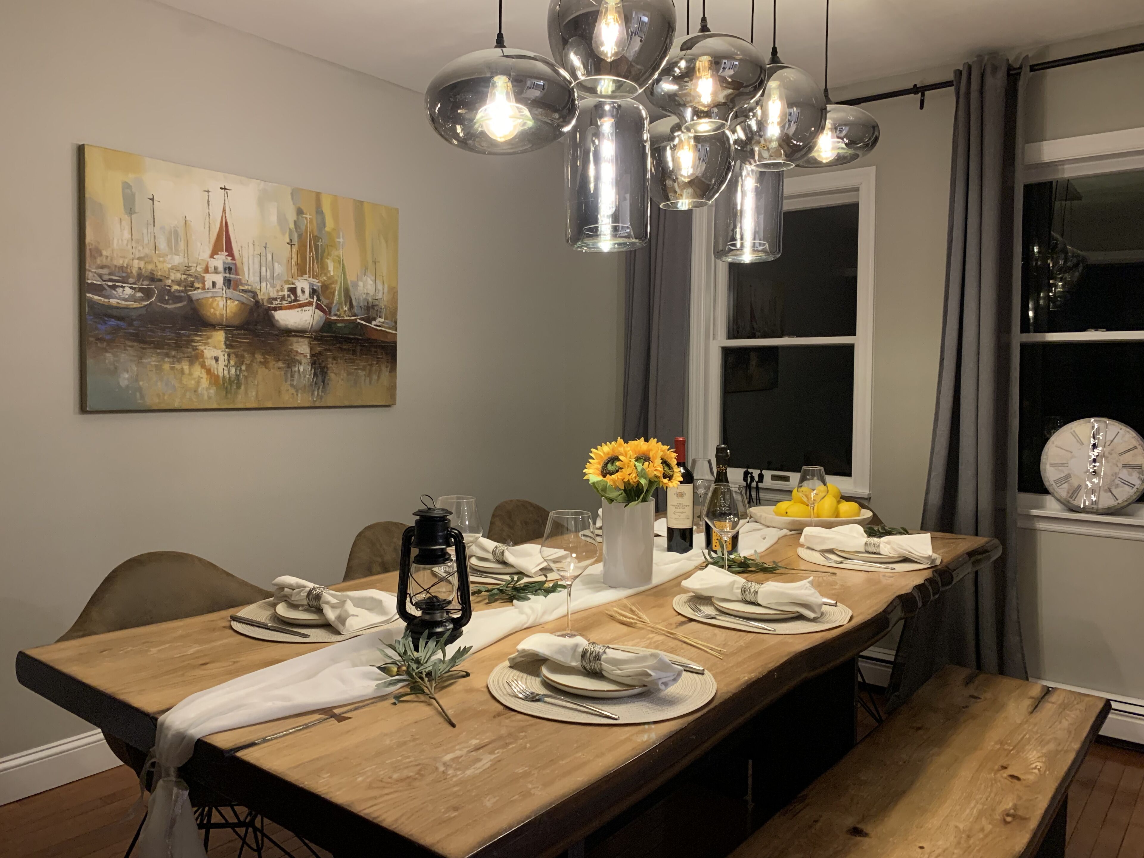 Un comedor con una mesa de madera dispuesta para seis personas, un cuadro de barcos en la pared y un reloj grande también en la pared.