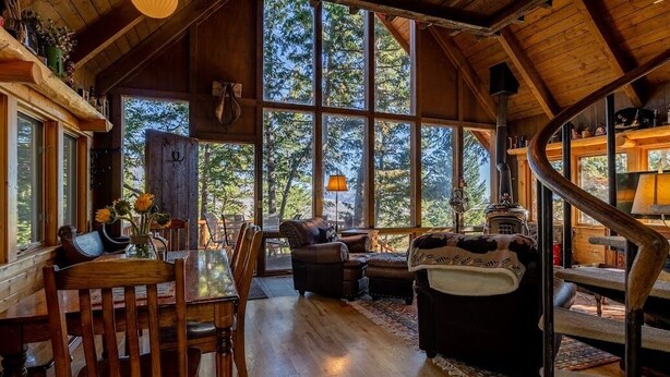 Interior acogedor de una cabaña con comedor, chimenea y grandes ventanales que dan al bosque.