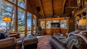 Interior rústico de una cabaña con muebles de cuero, chimenea y un ventanal con vistas a un bosque.