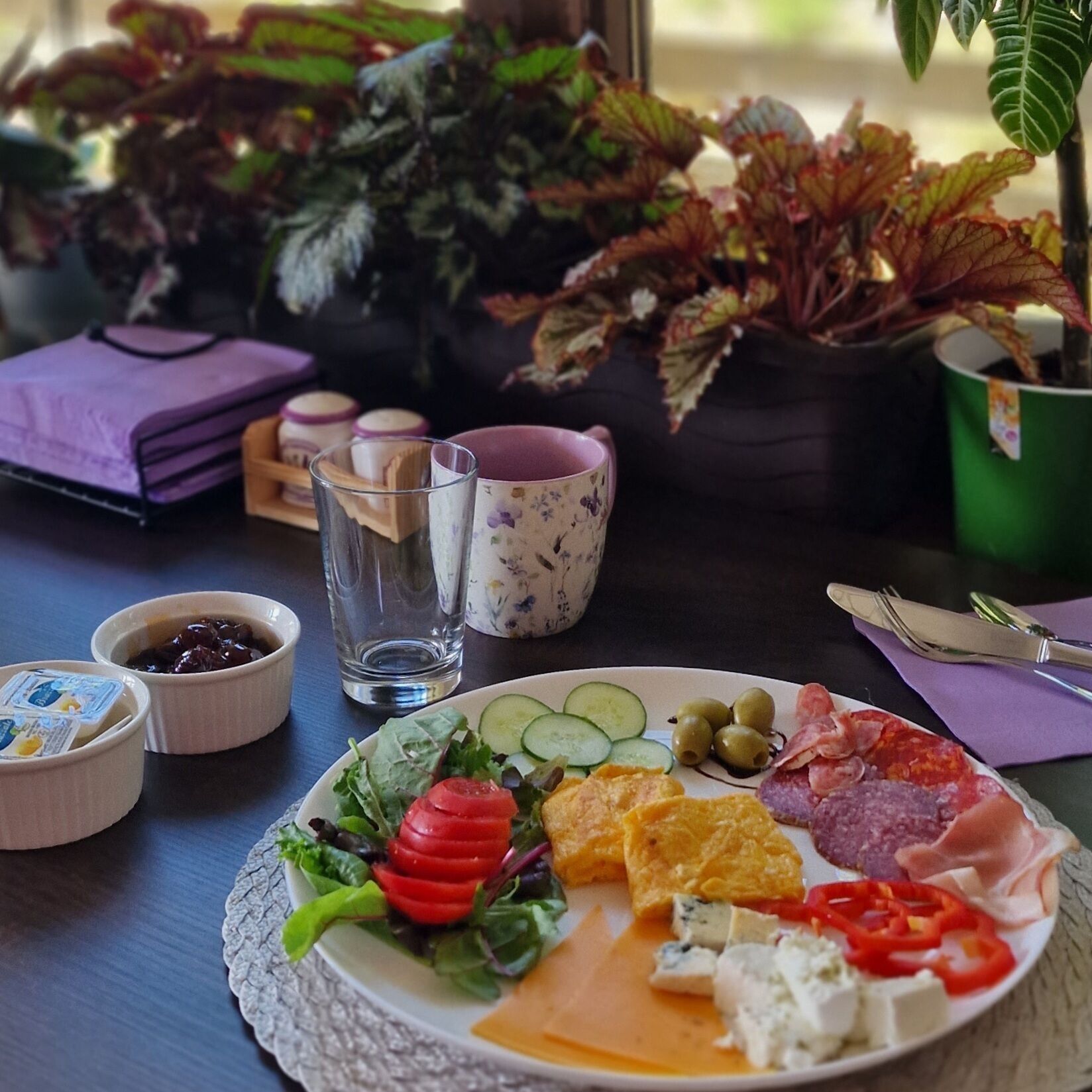 A table with a plate of assorted cheeses, cured meats, and vegetables, accompanied by small bowls of condiments and a glass of water.
