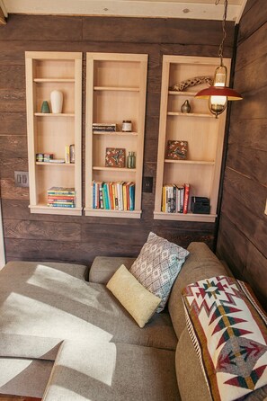 Um quarto aconchegante com um sofá, almofada decorativa estampada e estantes de madeira repletas de livros e objetos decorativos.