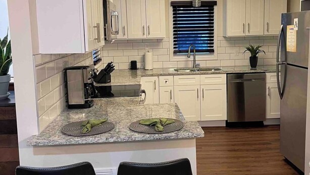 A modern kitchen with white cabinets, a granite countertop, and a stainless steel refrigerator. There are two bar stools and a window with blinds. - Blue Bungalow-Downtown Acworth (Acworth)