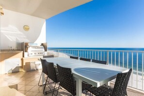 Un balcon avec une table et des chaises blanches, un barbecue et une vue sur la mer.