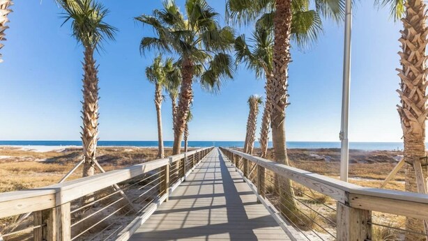 En träpromenad med räcken, palmer och utsikt över stranden och havet.