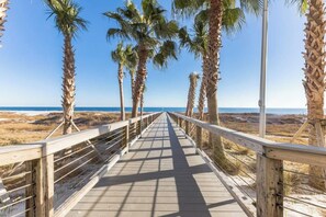 En träpromenad med räcken, palmer och utsikt över stranden och havet.