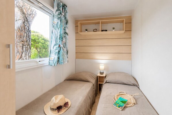 Um quarto pequeno e acolhedor com duas camas de solteiro, uma mesa de cabeceira com abajur, uma prateleira sobre as camas e uma janela com cortinas.