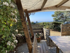 A wooden deck with a pergola overhead, surrounded by greenery and a view of the sea.