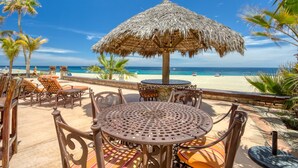 Een terras aan het strand met een rieten parasol, rieten meubels en uitzicht op de oceaan.