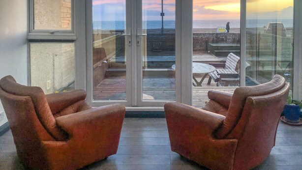 Un balcon avec deux fauteuils en cuir, une table en bois et une vue sur la mer et le coucher de soleil.
