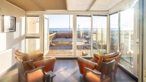 Un salon moderne avec des fauteuils en cuir et une petite table, de grandes fenêtres offrant une vue sur la mer, et un balcon aménagé avec du mobilier d’extérieur.
