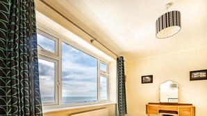 Une chambre avec une grande fenêtre, une commode en bois, un miroir et un luminaire suspendu.