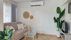 Ein Wohnzimmer mit einem beigefarbenen Sofa, einem Glastisch, einer TV-Konsole und einer großen Topfpflanze.