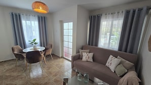 Ein Wohnzimmer mit einem braunen Sofa, einem Glastisch, einem Esstisch mit Stühlen und einer hängenden Pendelleuchte.