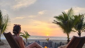 A person relaxing on a beach chair with a sunset view, a fire pit, and two bottles on a wooden table.