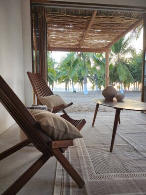 A beachside lounge with wooden lounge chairs and a table, overlooking a palm tree-lined beach.
