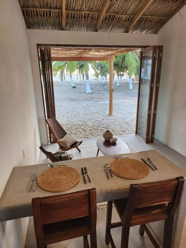 A dining area with a table set for two, a chair, and a view of a sandy beach with palm trees.
