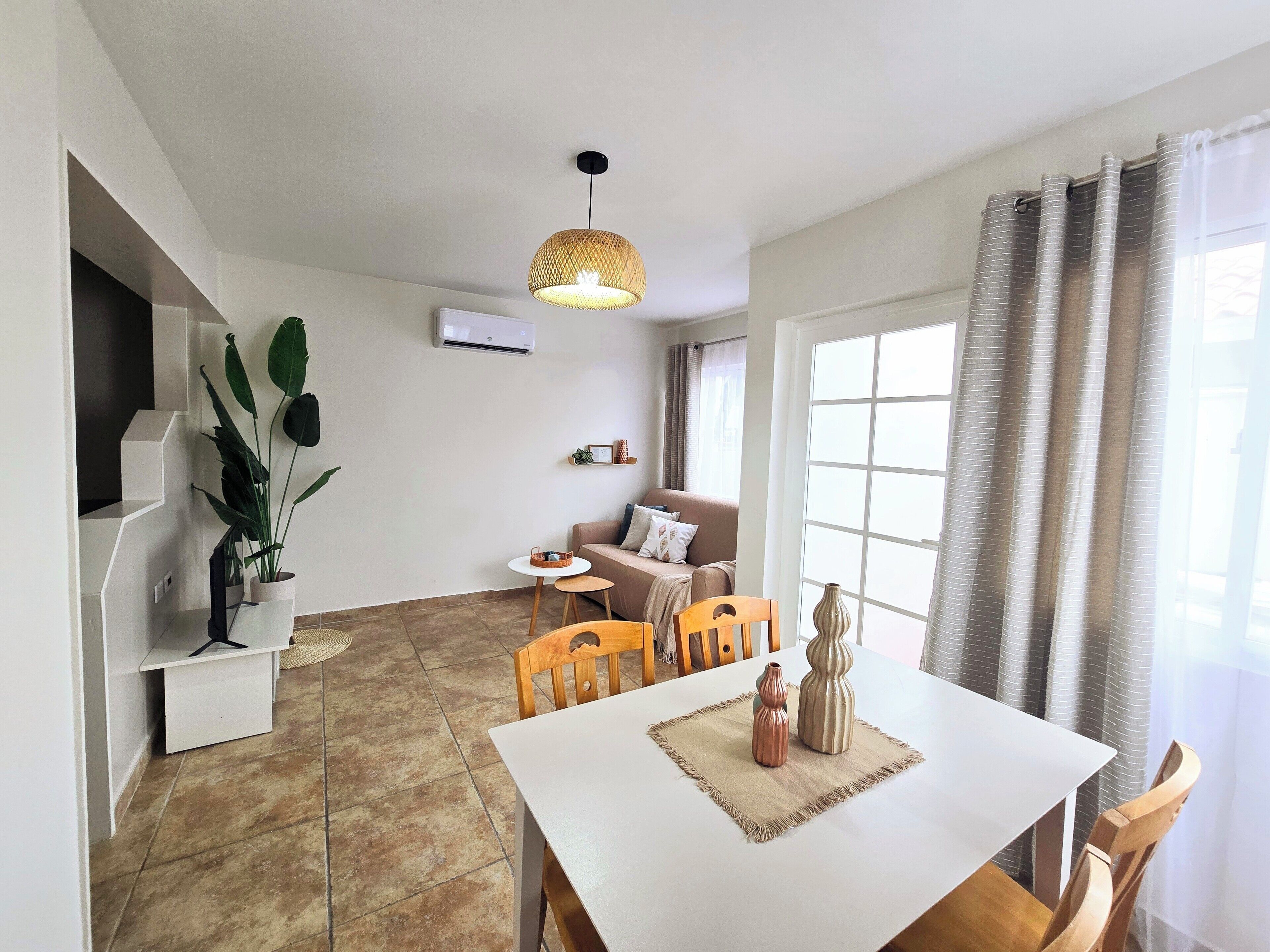 A modern living room with a dining area, featuring a white table, wooden chairs, a hanging light fixture, and a sofa.