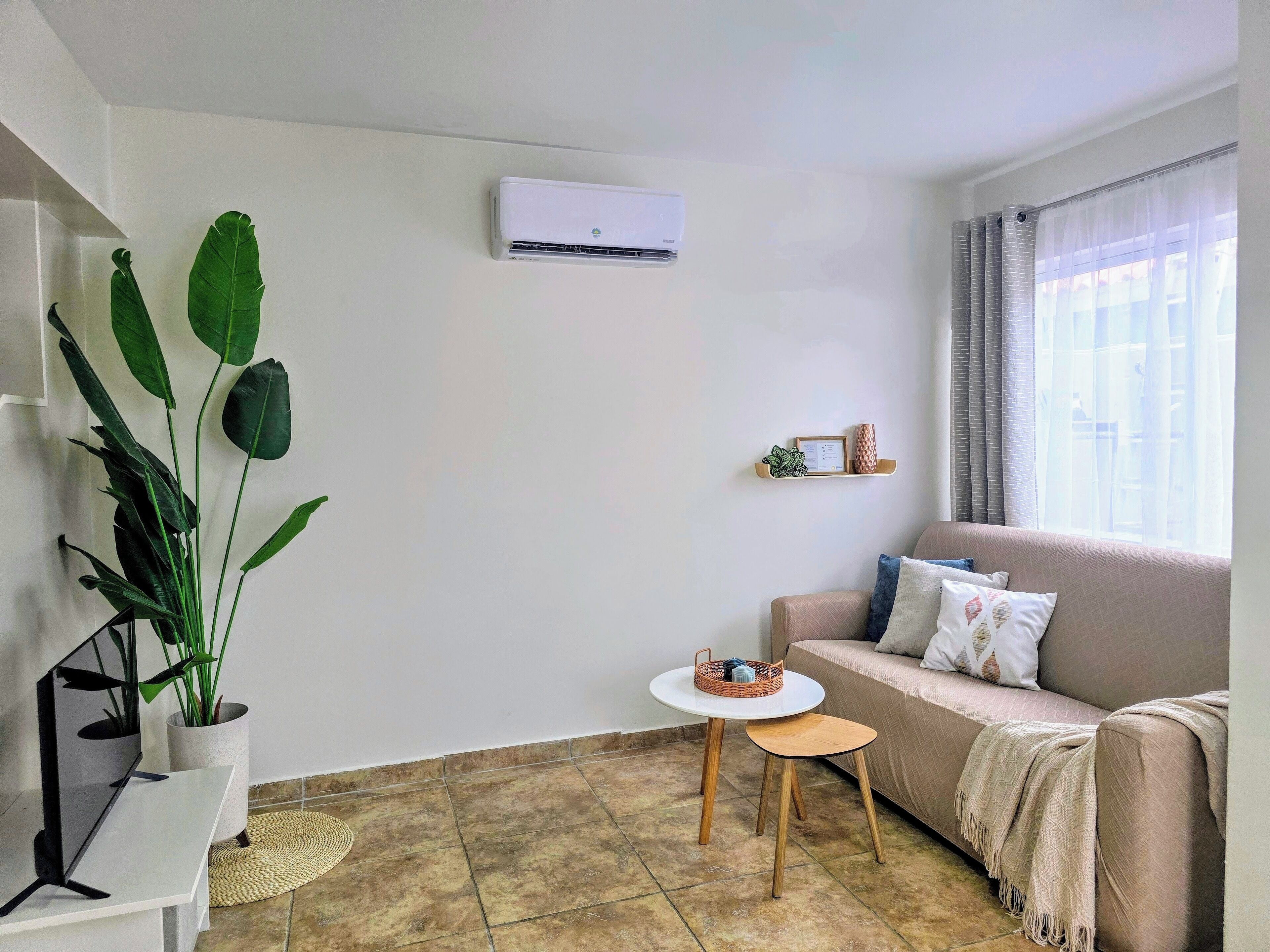 Un salón con un sofá color beige, una mesita redonda, una planta en maceta y un aire acondicionado instalado en la pared.