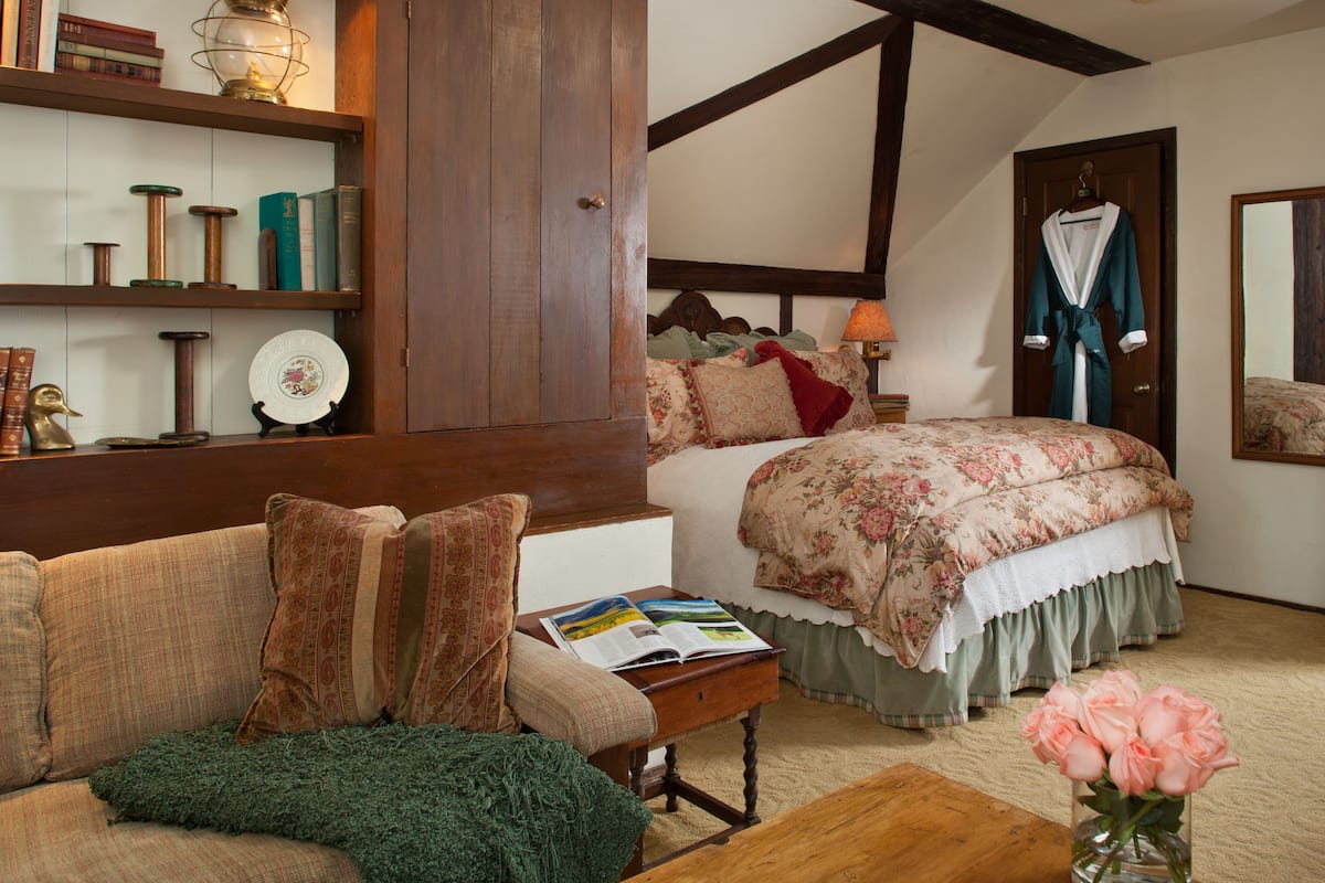 Um quarto com uma cama, um guarda-roupa de madeira, uma mesa de cabeceira com um livro, um cobertor verde no sofá e um vaso com flores.