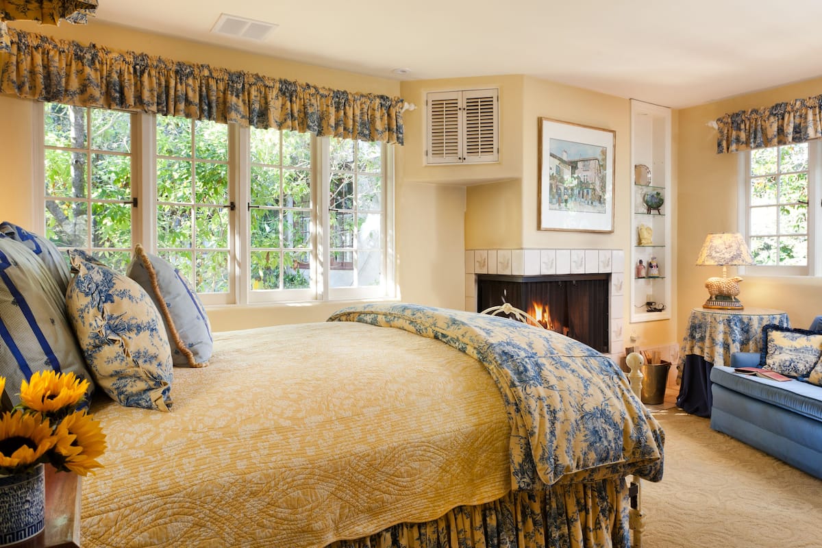 Quarto com uma cama grande, lareira, sofá azul e janela com vista para o verde.