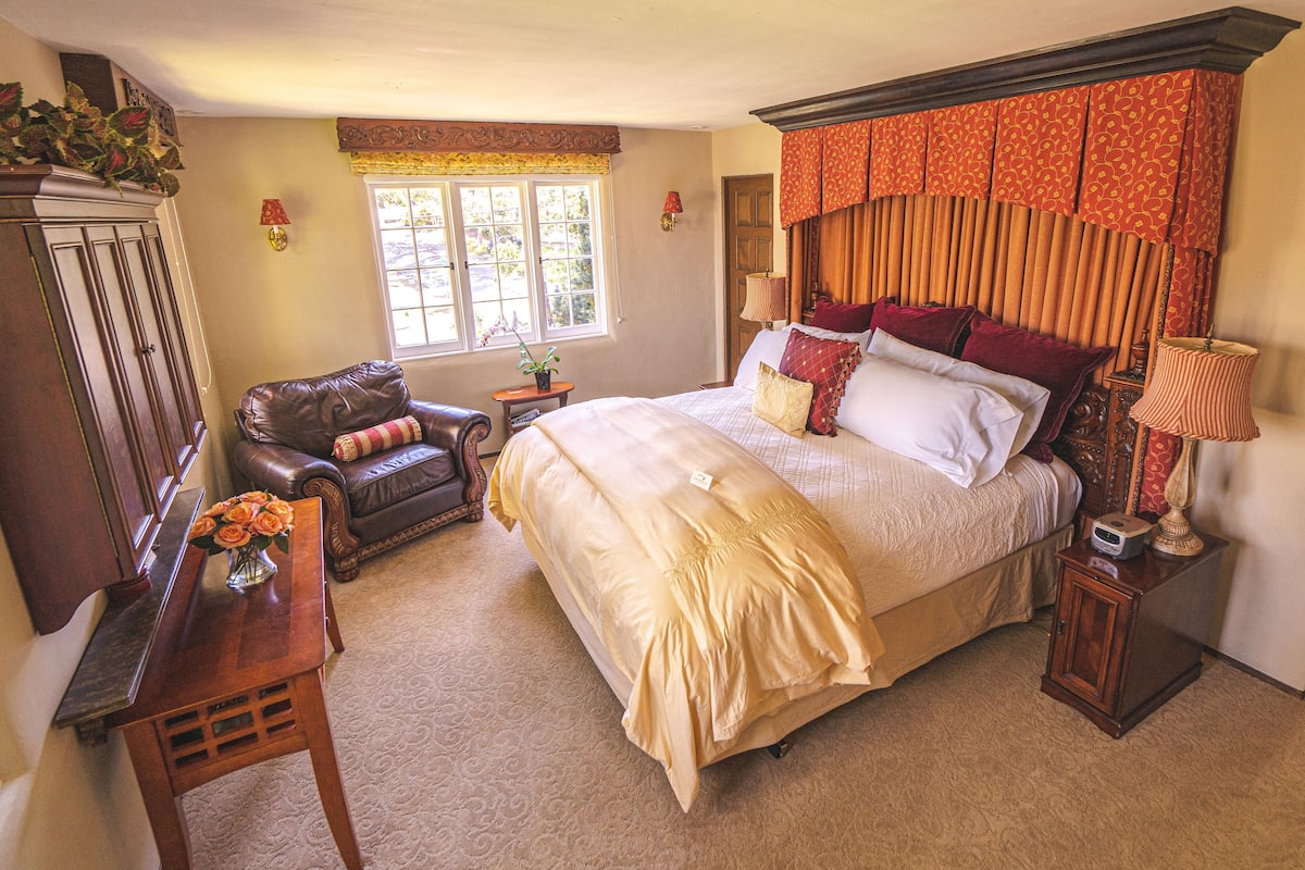 Quarto com uma cama grande, mesa de cabeceira, luminária, cadeira e escrivaninha com um vaso de flores.