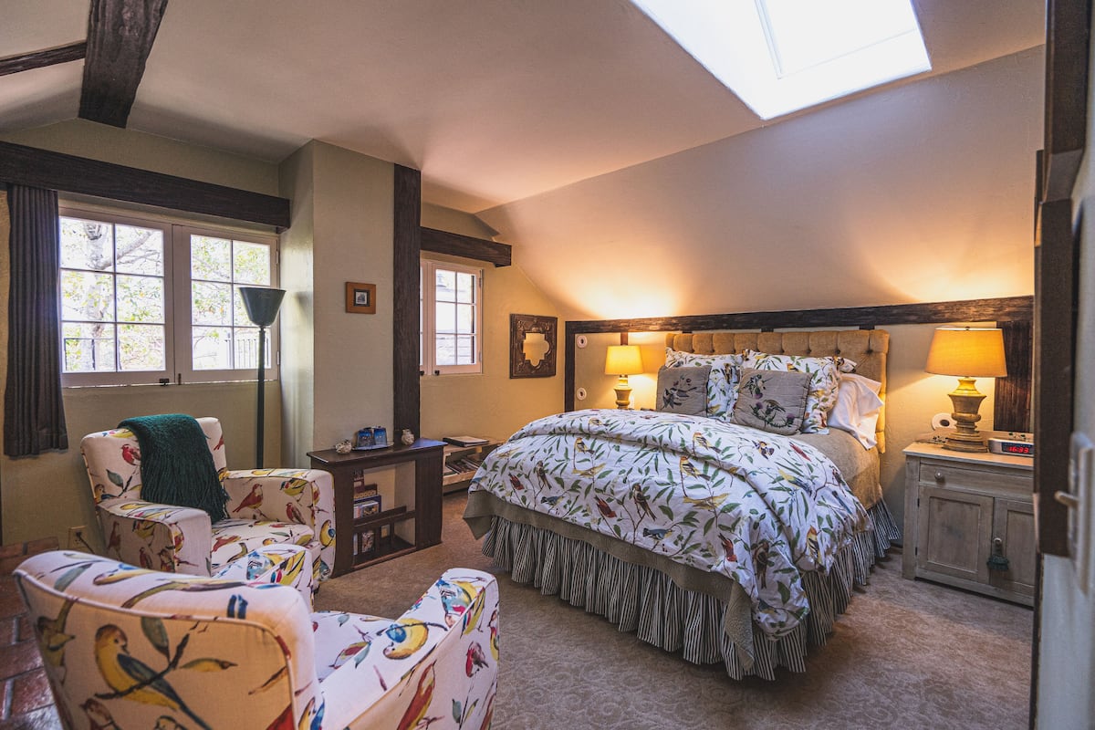 Quarto com cama grande, duas cadeiras, mesa de cabeceira e claraboia.
