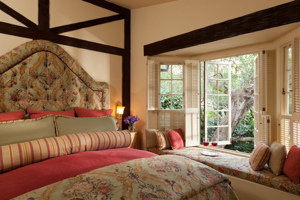 Quarto com uma cama grande, um banco com almofadas e uma janela com persianas.