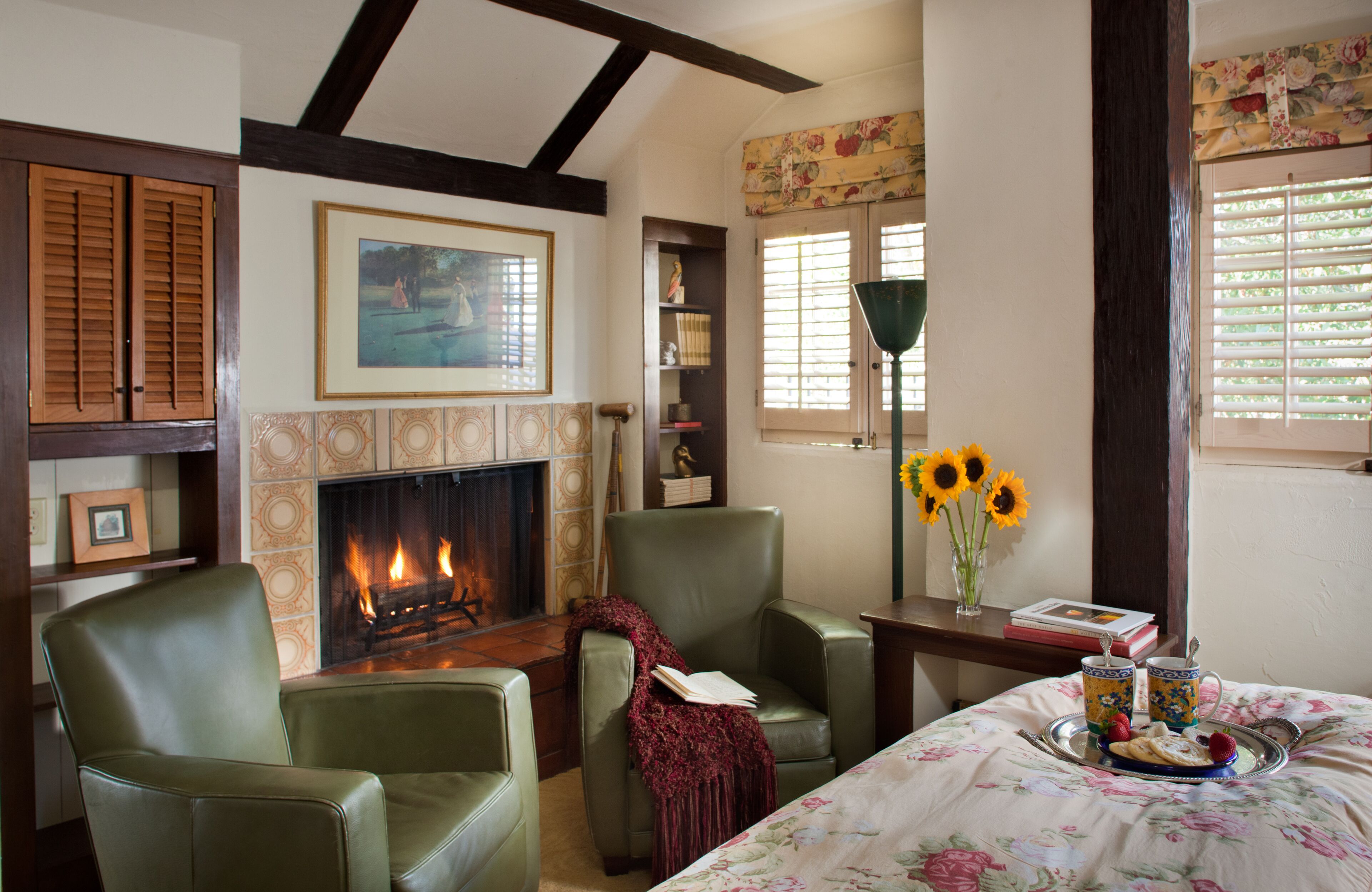 A cozy living room with a fireplace, green leather chairs, a floral bedspread, and a tray with food and drinks.