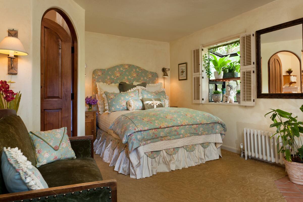 Um quarto com colcha florida, porta de madeira, mesa de cabeceira com abajur, janela com persianas e radiador.