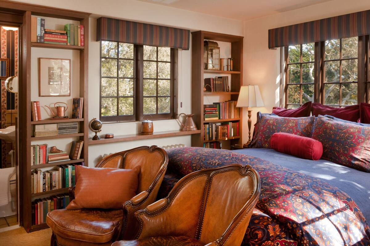 Um quarto acolhedor com uma cama grande, uma cadeira de madeira e estantes repletas de livros e objetos decorativos.